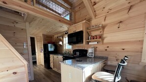 Interior de una cabaña de madera con zona de cocina, comedor y una escalera que sube a un nivel superior.