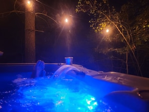 A hot tub with a person lying down, surrounded by trees and string lights.