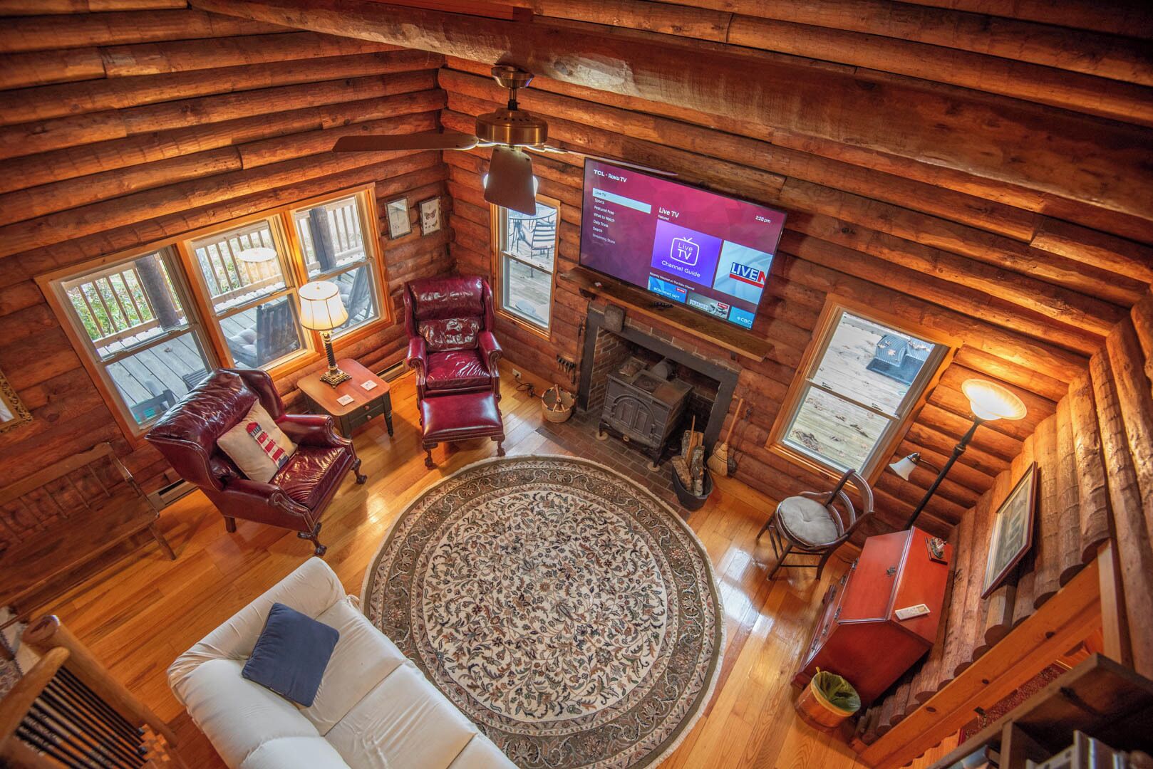 A log cabin interior with a fireplace, a TV mounted on the wall, and a cozy seating area with a patterned rug.