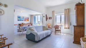 A spacious living room with a sofa, dining table, and chairs. There is a large window with curtains, a lamp, and a wooden cabinet. - Cosy apartment for 4 guests with WIFI, A/C, TV, patio and pets allowed (San Vincenzo)