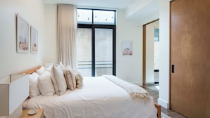 Een slaapkamer met een groot bed, een nachtkastje, een raam met gordijnen en een deur naar een andere kamer.