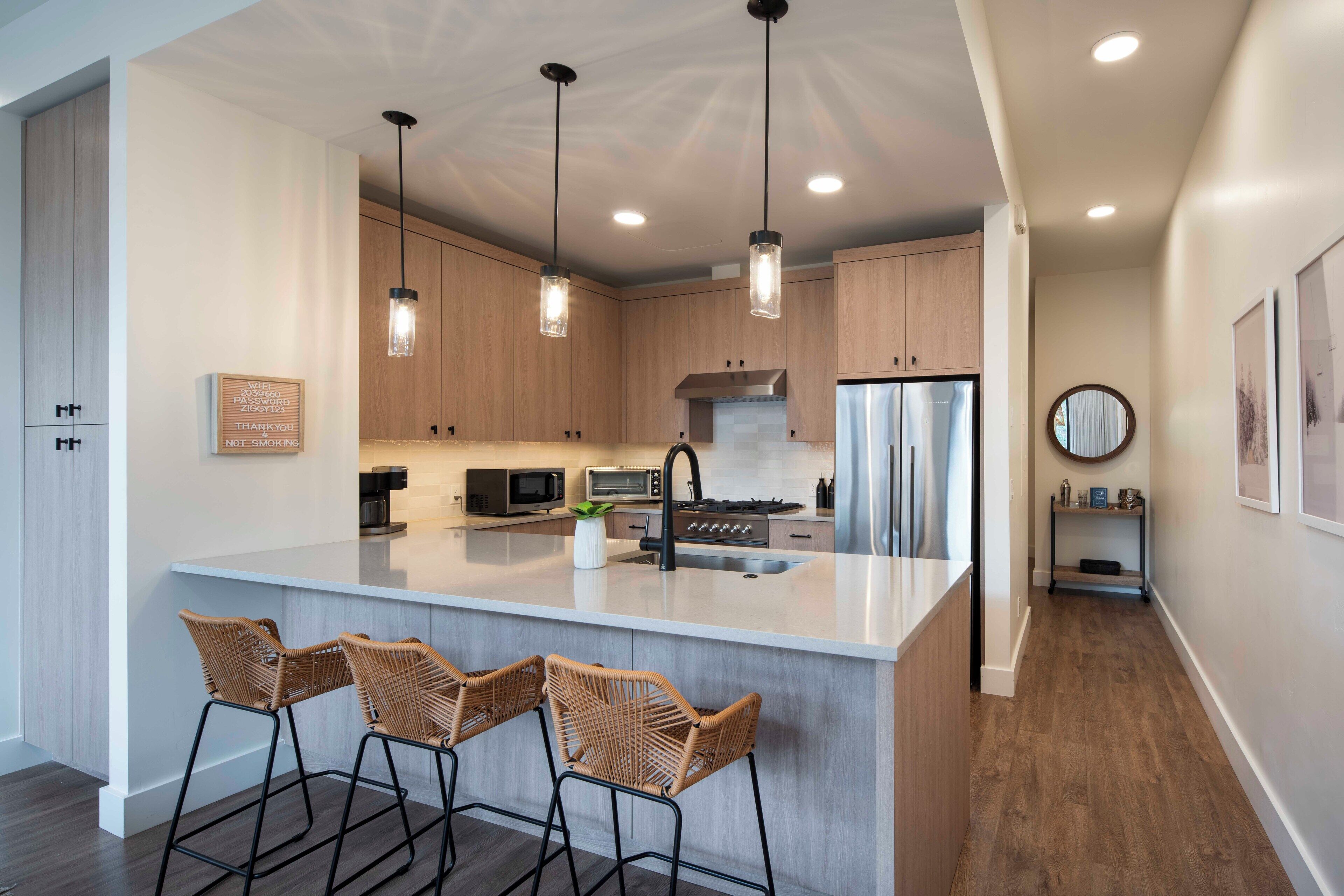 A modern kitchen with a large island, bar stools, pendant lighting, and stainless steel appliances.