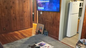 Uma sala de estar com paredes de madeira, uma televisão presa na parede, uma mesinha com uma revista e uma porta de entrada para uma cozinha.