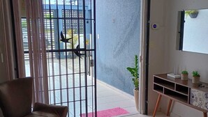 Een woonkamer met een schuifpui die uitkomt op een balkon, een bruine leren bank, een gestreept fauteuil, een klein houten bijzettafeltje met planten en een aan de muur bevestigde televisie.
