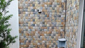 Een kleine, afgesloten douchecabine met een betegelde wand, een handdouche en een klein plankje.