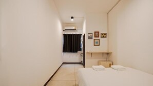 A hotel room with a bed, a small bedside table, a wall-mounted shelf, and a window with curtains. - GO INN Asiatique (Bangkok)