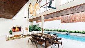 A spacious indoor area with a dining table set for six, surrounded by chairs, a large TV screen displaying a fire, and a pool in the background. - Luxury Noah Villa (Kuta)