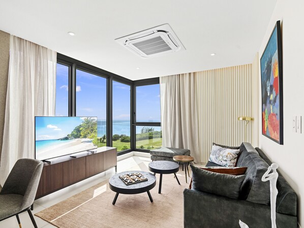 A modern living room with a large TV, a sofa, a coffee table, and a view of the ocean. - B-701 Stunning corner ocean view (Cupecoy)