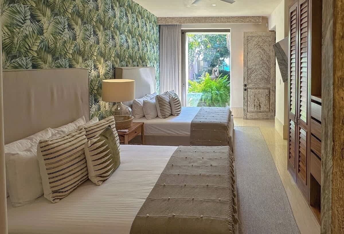 A bedroom with a bed, pillows, a nightstand, and a lamp. There is a view of greenery outside the window.