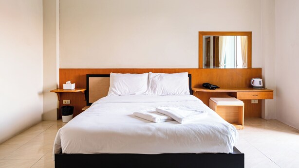 Un lit bien fait, avec du linge de lit blanc, une tête de lit en bois, une table de chevet avec une lampe et une petite table de nuit en bois avec une poubelle.