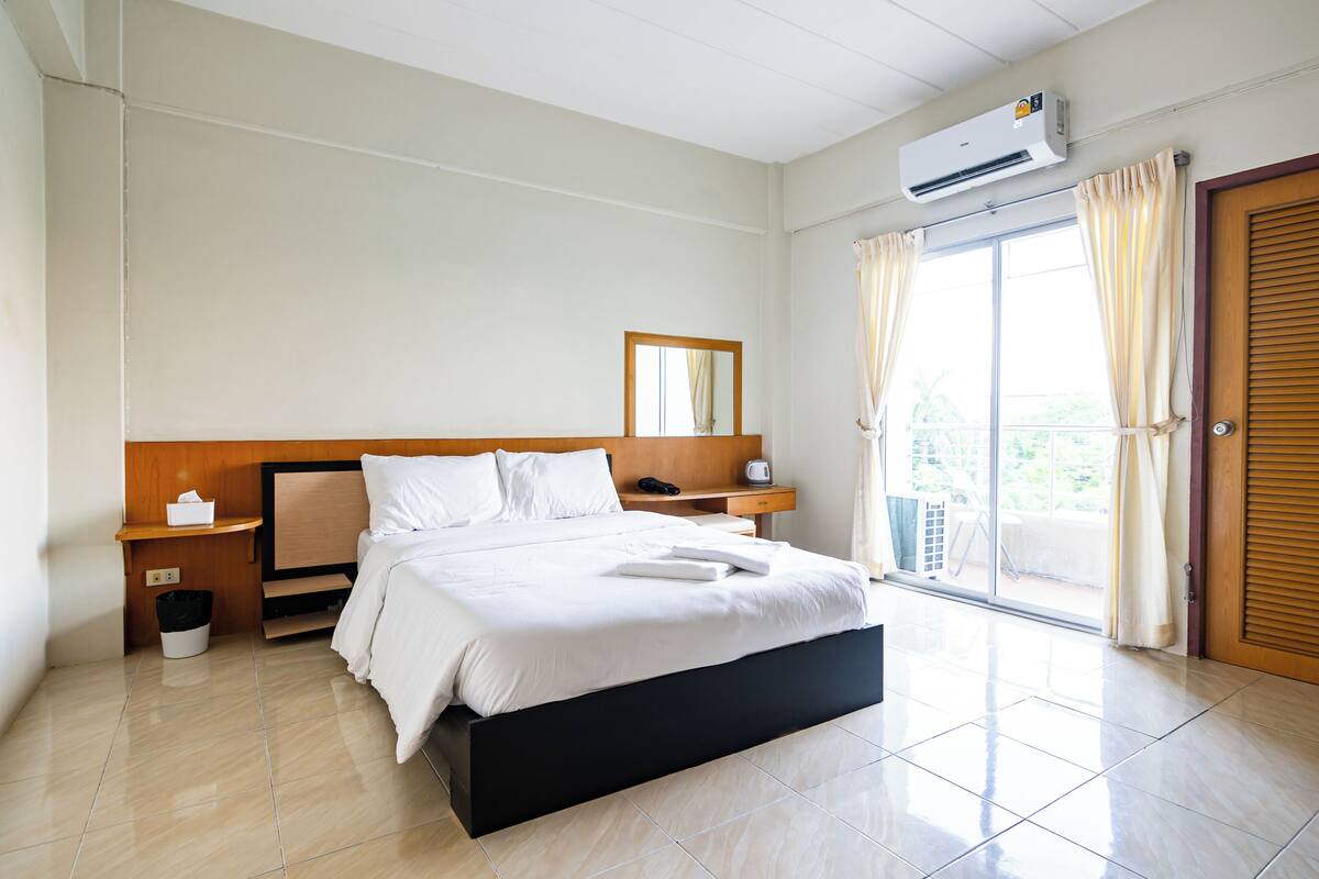 A hotel room with a bed, bedside tables, a mirror, an air conditioning unit, a window with curtains, and a door.
