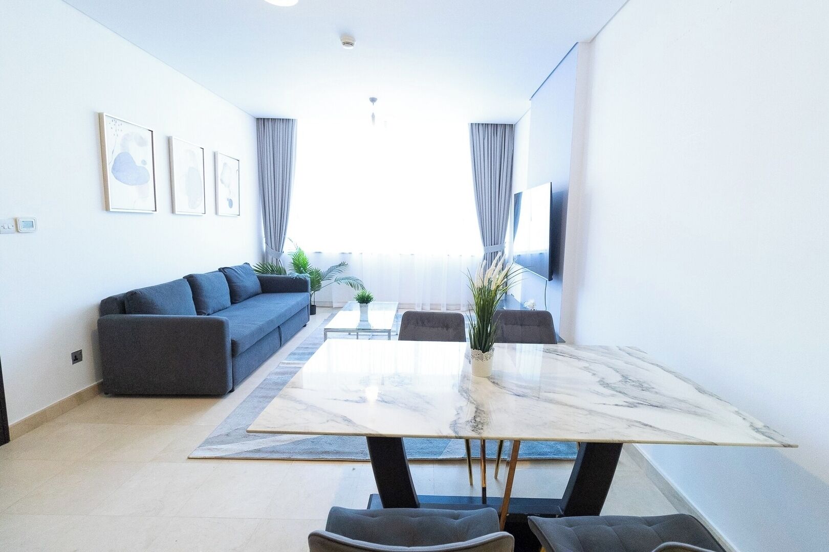 A modern living room with a sofa, a dining table, and chairs. There are curtains, a plant, and a TV on the wall.
