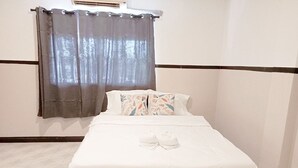 A neatly made bed with white linens and two pillows, a gray curtain partially drawn, and a white wall with a dark trim. - Mitr inn Phuket Old town (Phuket)