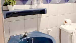 A bathroom with a blue sink, a mirror, a toilet, and a shelf with toiletries. - Mitr inn Phuket Old town (Phuket)