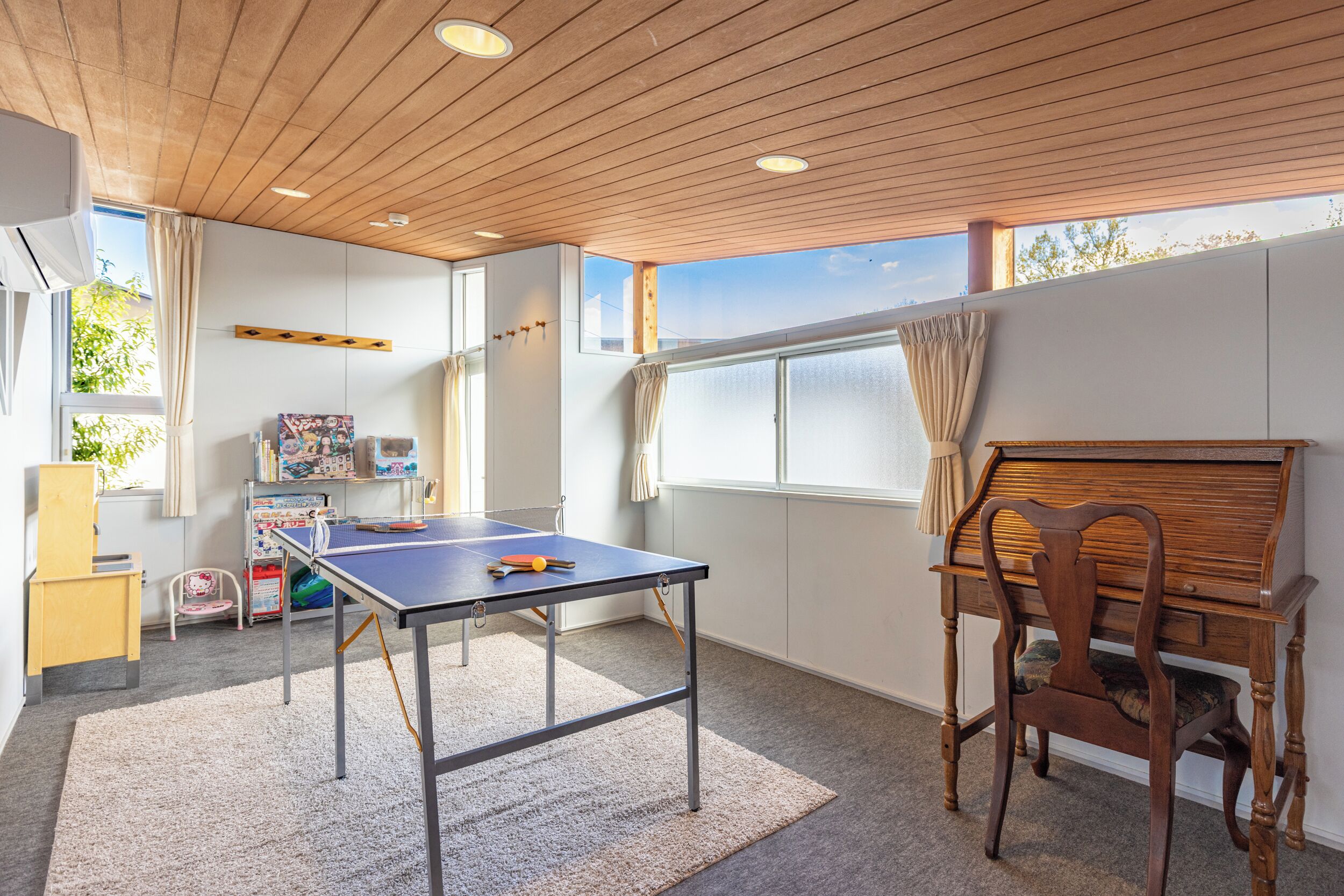 A room with a ping pong table, a wooden chair, a bookshelf, and a window with curtains.