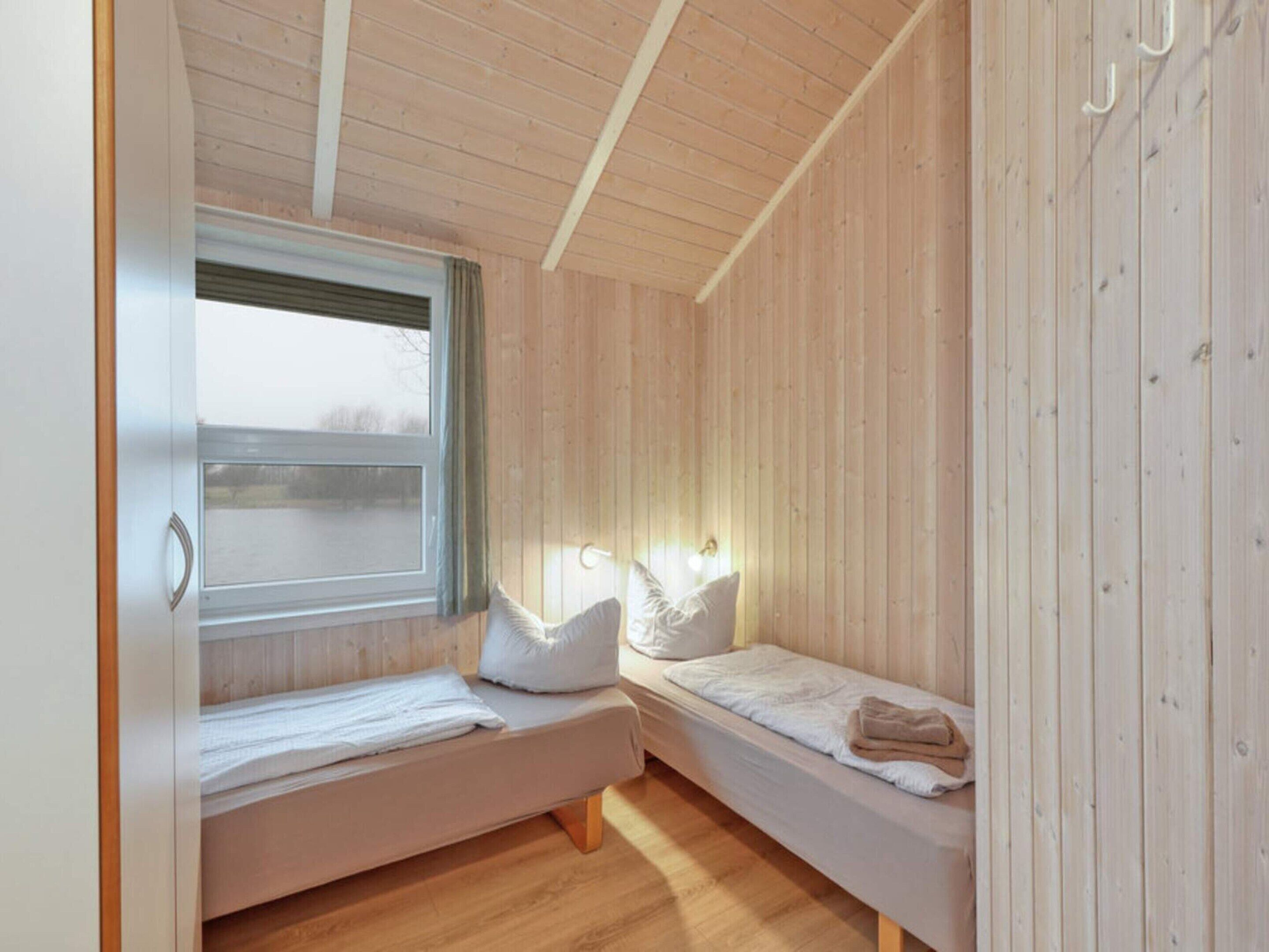 Quarto pequeno e acolhedor com duas camas de solteiro, paredes de madeira, uma janela com cortinas e teto de madeira.
