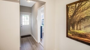 A hallway with a white door, a framed picture of a forest, and a blue chair in the background. - Chic & Cozy 3BR 2BA Home - Heart of GNV! (Gainesville)