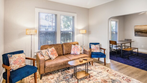 A living room with a brown leather sofa, two blue armchairs, a coffee table, and a patterned rug. There are two floor lamps, a dining table with chairs, and a painting on the wall. - Chic & Cozy 3BR 2BA Home - Heart of GNV! (Gainesville)