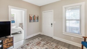 A bedroom with a bed, a TV, a dresser, a rug, a door, and a window with blinds. - Chic & Cozy 3BR 2BA Home - Heart of GNV! (Gainesville)