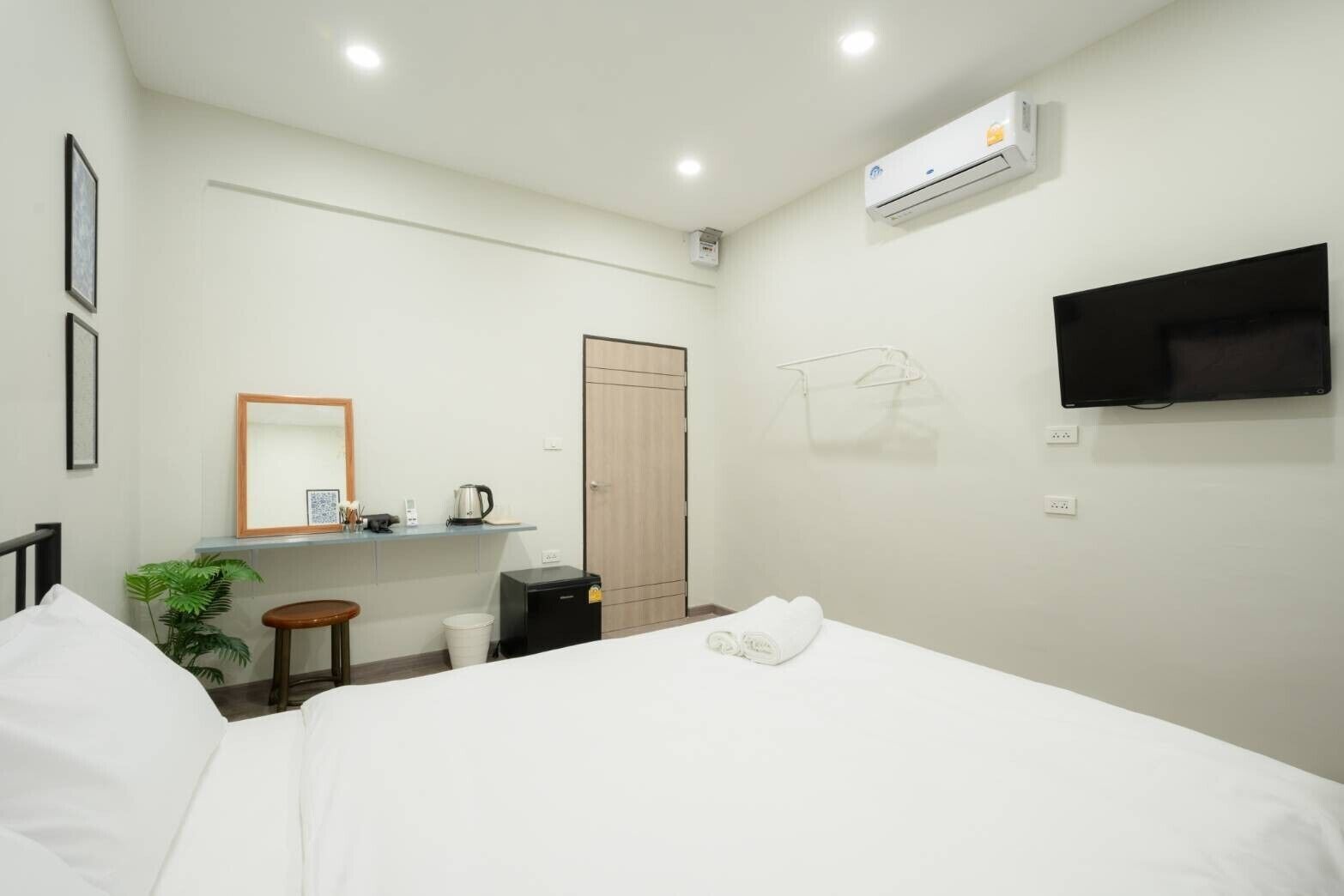 A hotel room with a bed, a desk with a kettle, a small trash bin, a plant, and a TV mounted on the wall.