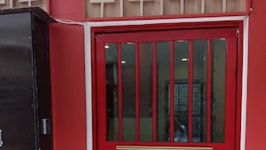 A red door with a metal security bar, a black cabinet to the left, and patterned tiles on the floor. - CASA Yaowarat (Bangkok)