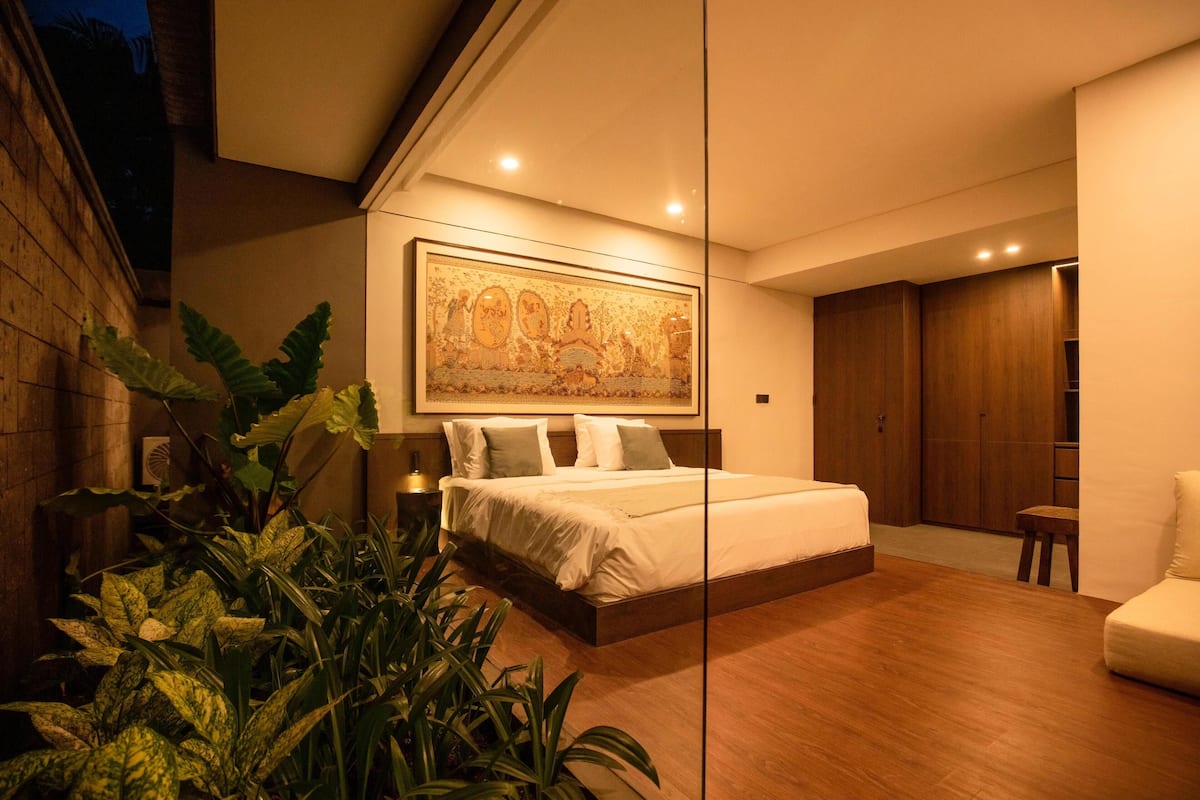 A modern bedroom with a large bed, a wooden floor, a large plant, and a painting on the wall.