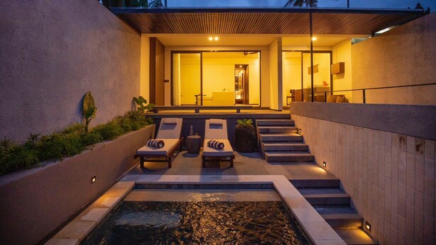 Un área de piscina moderna al aire libre, con sillones reclinables, una pequeña cascada y una escalera que conduce a una entrada cubierta.