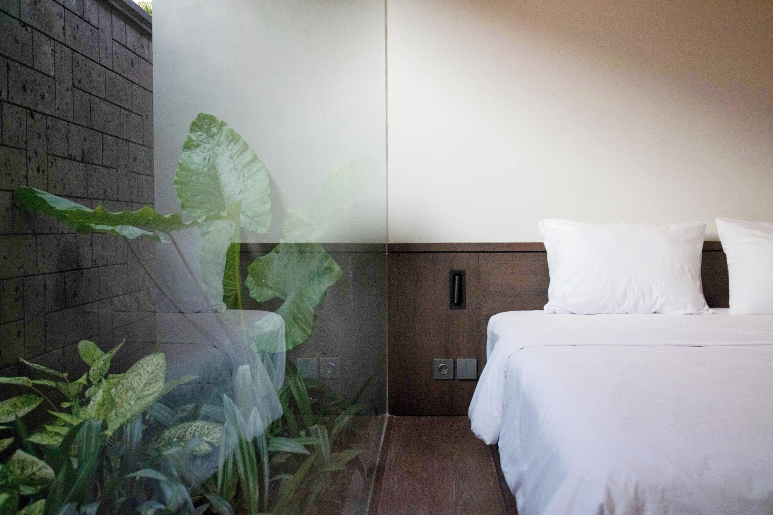 A modern hotel room with a large bed, a wooden headboard, and a view of a brick wall and greenery outside.