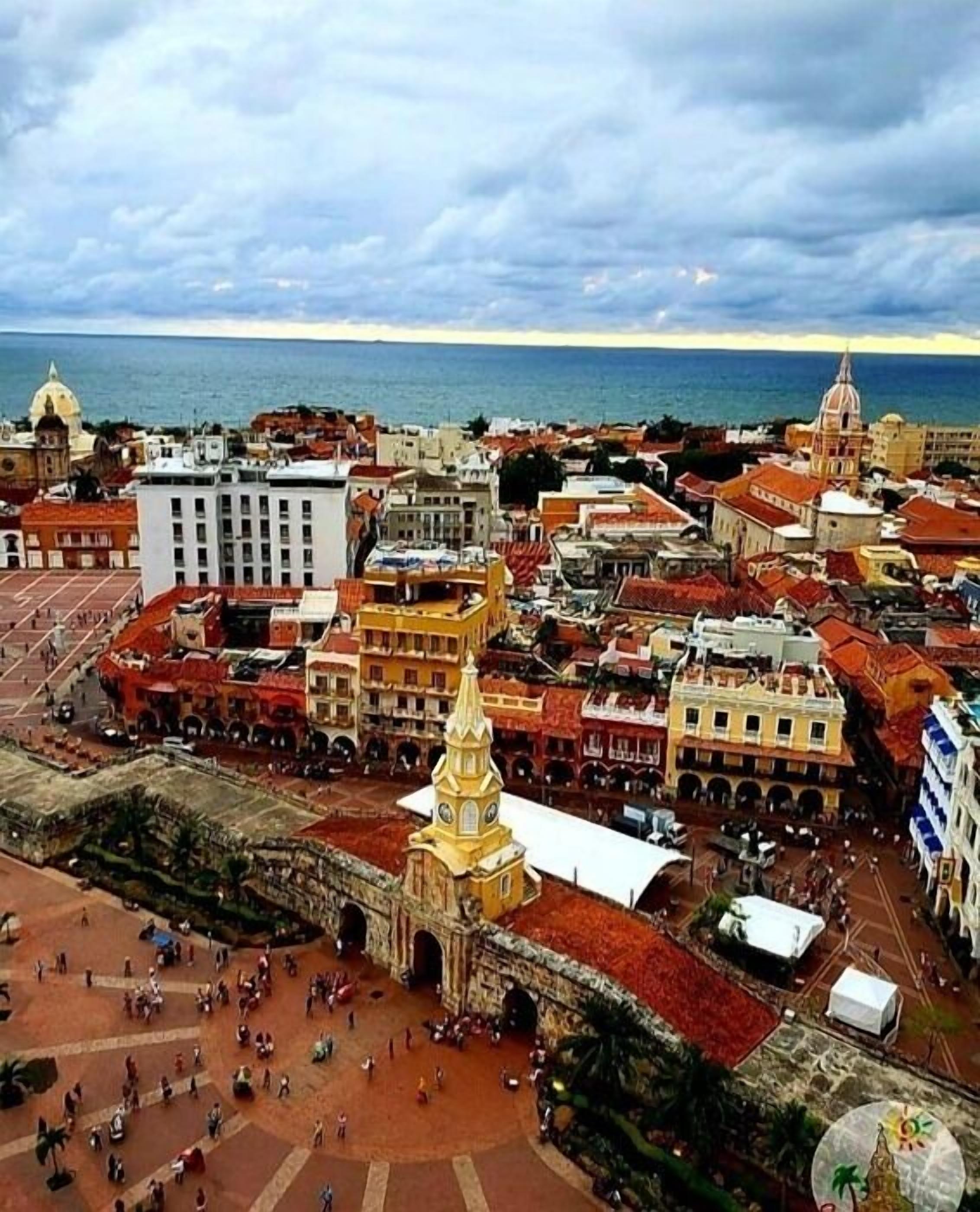 An aerial view of a historic city with a prominent church featuring a tall bell tower, surrounded by colorful buildings and a large open square.