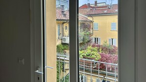 Une vue à travers la fenêtre d’un bâtiment à la façade jaune, doté d’un toit recouvert de tuiles rouges et d’un balcon orné de plantes en pot.