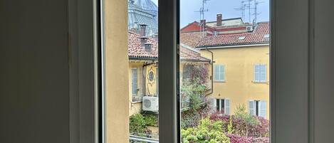 A view through a window of a building with a yellow facade, a red-tiled roof, and a balcony with potted plants.