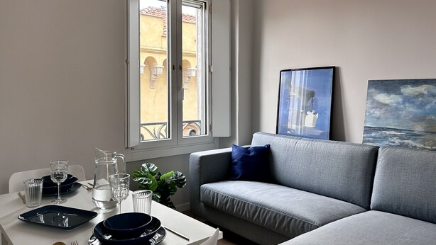 Un salon moderne avec un canapé gris, une table à manger dressée avec des assiettes, des verres et des couverts, et une fenêtre donnant sur des immeubles.