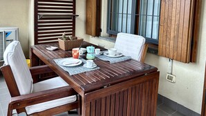 Uma mesa de jantar de madeira posta com pratos, chávenas e uma cafeteira, com uma janela e persianas ao fundo.