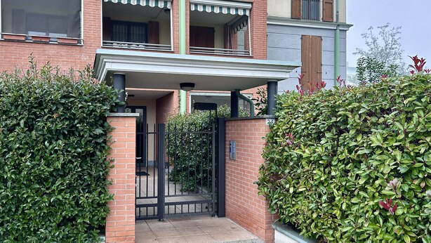 Um edifício residencial de vários andares com fachada de tijolos, uma varanda com plantas em vasos e uma entrada com portão.