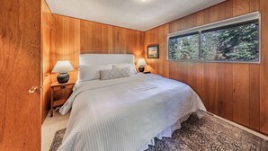 A bedroom with wooden panel walls, a large bed with white bedding, two bedside lamps, a window with a forest view, and a ceiling light. - Central 3 Bedroom House Less Than a Mile to Heavenly (South Lake Tahoe)