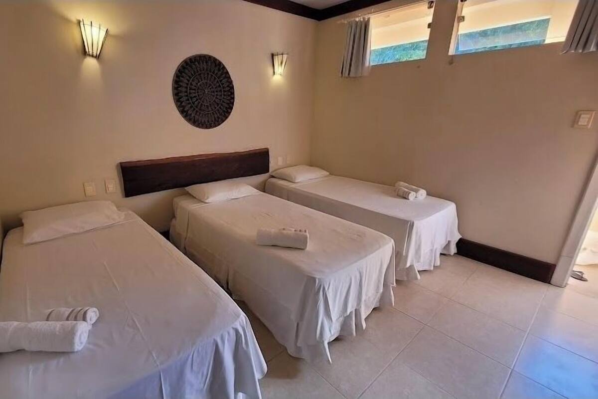 A hotel room with three single beds, each with a white bedspread and a rolled towel. There is a decorative wall piece above the beds and a window with curtains on the right wall.
