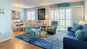 Un salon avec un canapé bleu, des fauteuils turquoise, un coin repas avec des chaises blanches et une grande fenêtre avec des stores.