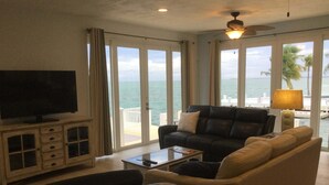 A living room with a television, sofa, and armchair, a ceiling fan, and a view of the ocean through large windows. - Open Ocean View, Private Hot Tub, Ice Machine, Moor 26' Boat! (Marathon)