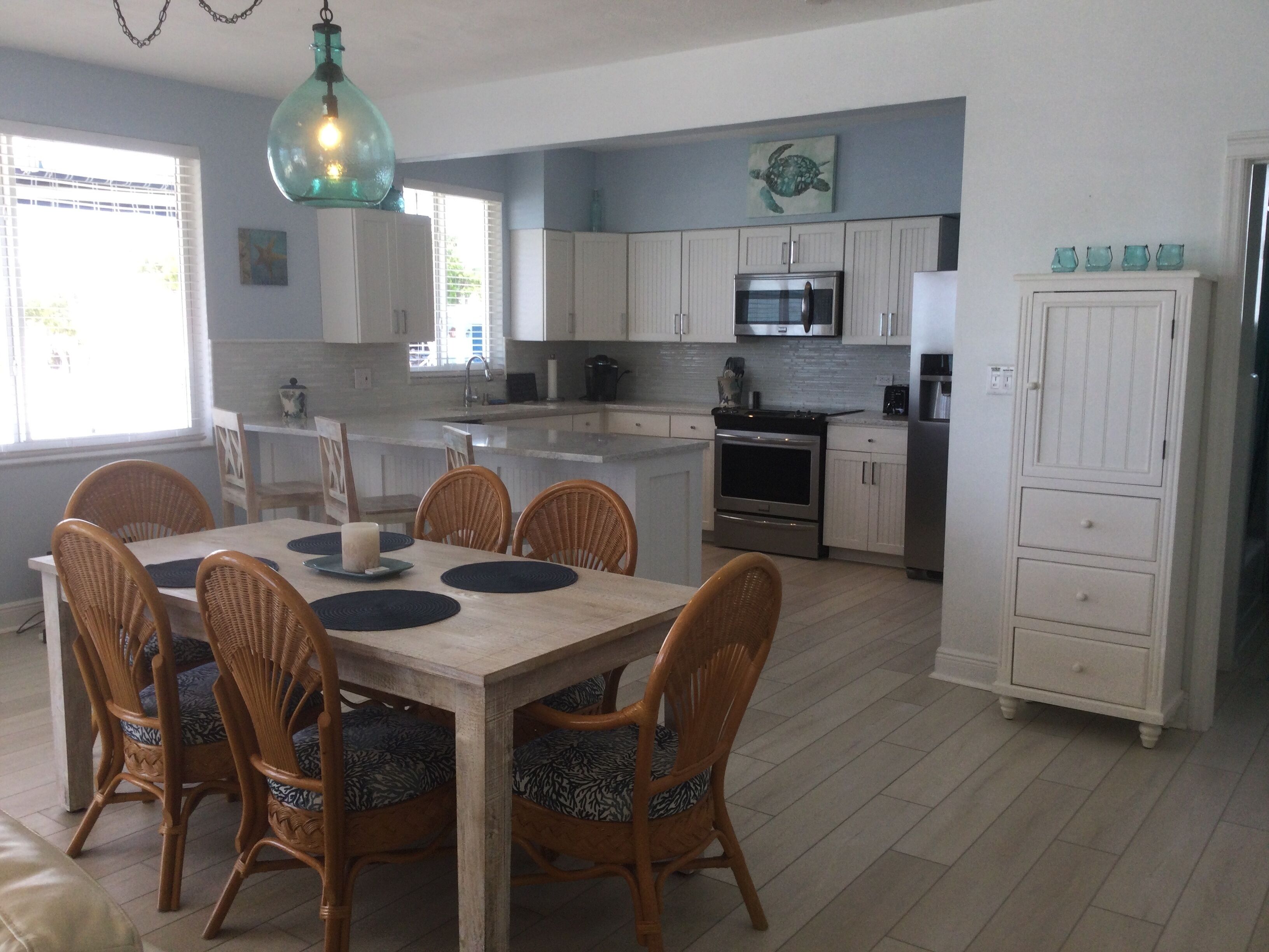 A modern kitchen with white cabinets, a central island, and a dining area with wicker chairs and a wooden table.