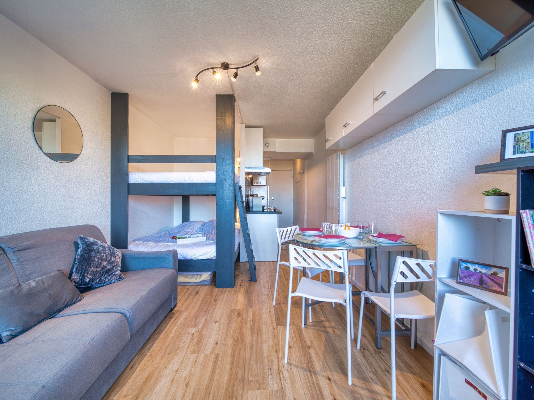 A compact living space with a bunk bed, a kitchenette, a dining area with white chairs, and a sofa with a cushion.
