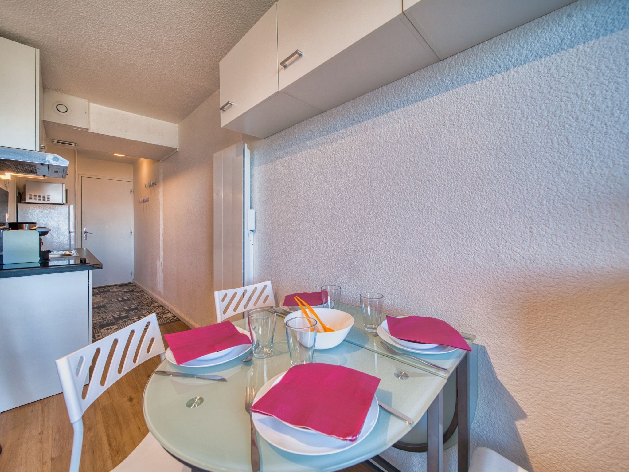 Zona pranzo con tavolo in vetro apparecchiata per quattro, sedie bianche e cucina sullo sfondo.