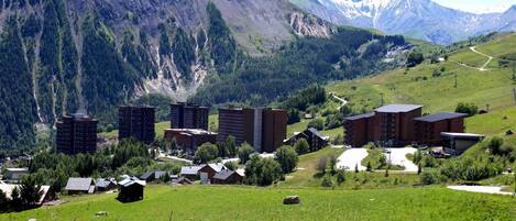 A mountainous landscape with a mix of modern and traditional buildings, including high-rise apartments and smaller houses, set against a backdrop of lush green fields and distant peaks.