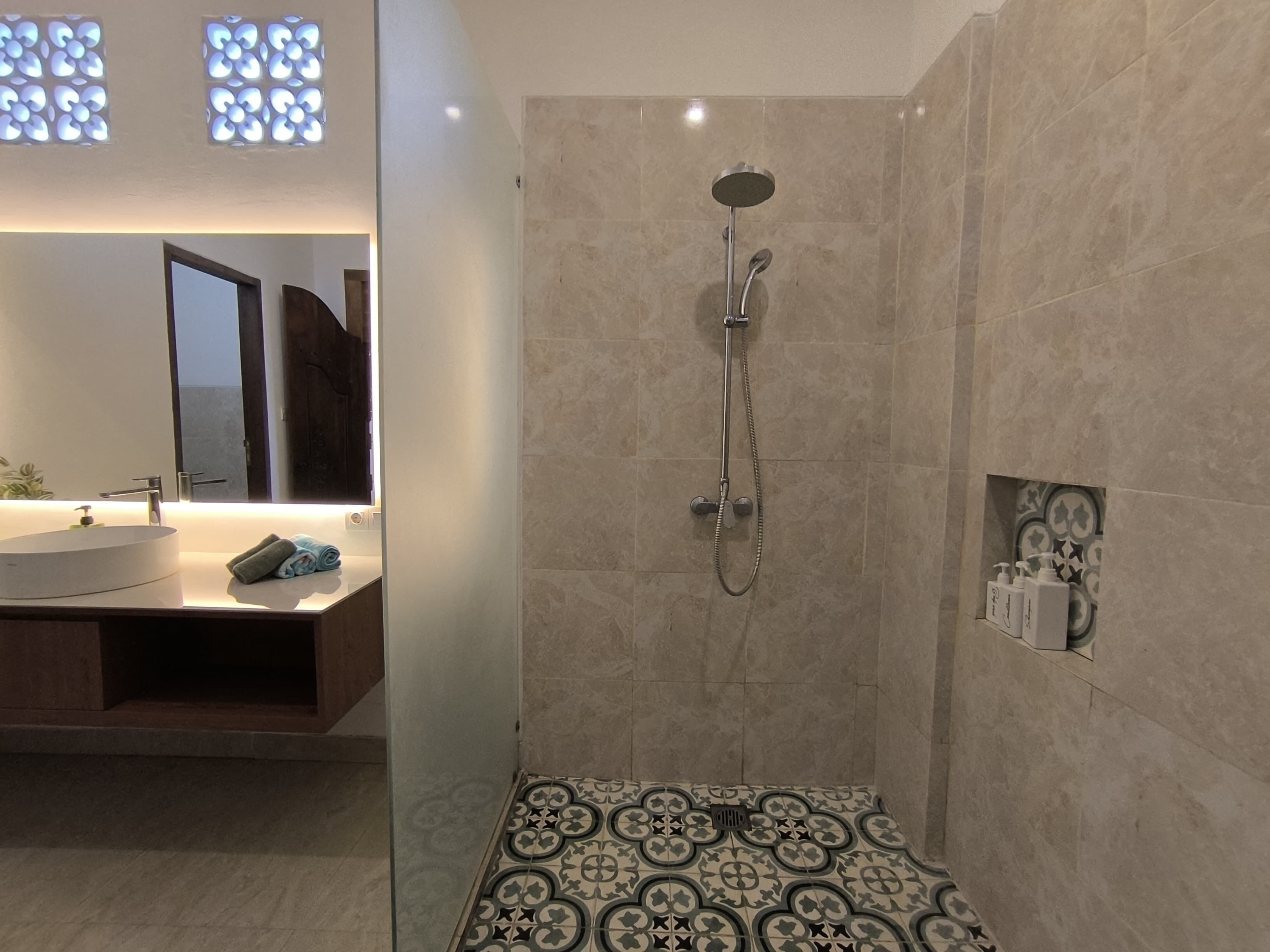 A modern bathroom with a large mirror, a sink, a shower with a handheld showerhead, and patterned floor tiles.