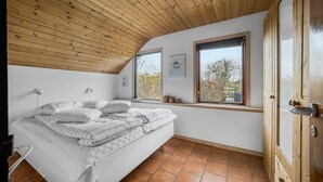 A bedroom with a sloped wooden ceiling, a bed with white bedding, two windows with artwork, and a wooden door.