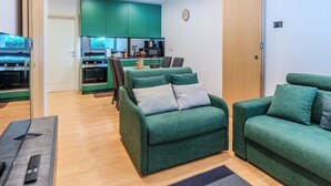 A modern living room with green sofas, a wooden dining table, and a kitchen in the background. - Rolando by Interhome (Lago di Mezzola)