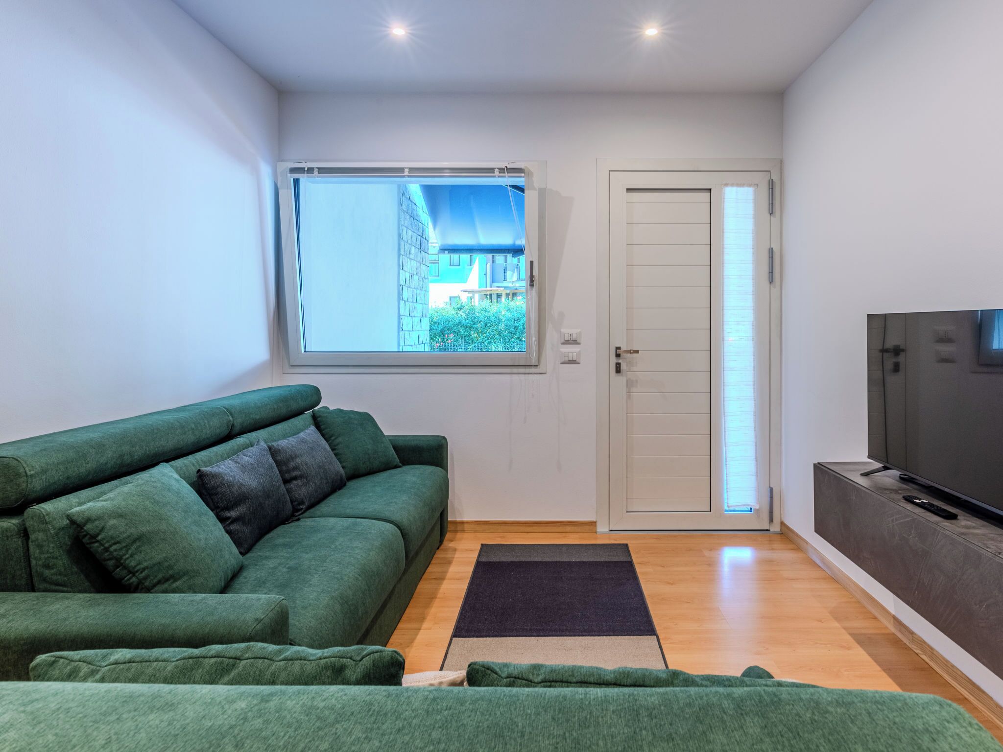 Een moderne woonkamer met een groene bank, een flatscreen-tv en een raam met uitzicht op gebouwen en groen.