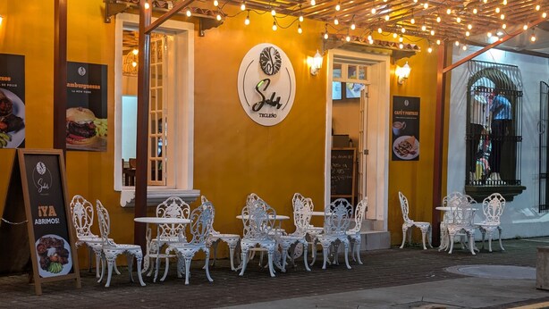 Un cafè a l'aire lliure amb caires i taules de metall blanc, una pèrgola de fusta amb llums penjades, i un rètol amb logotip i plats del menú.