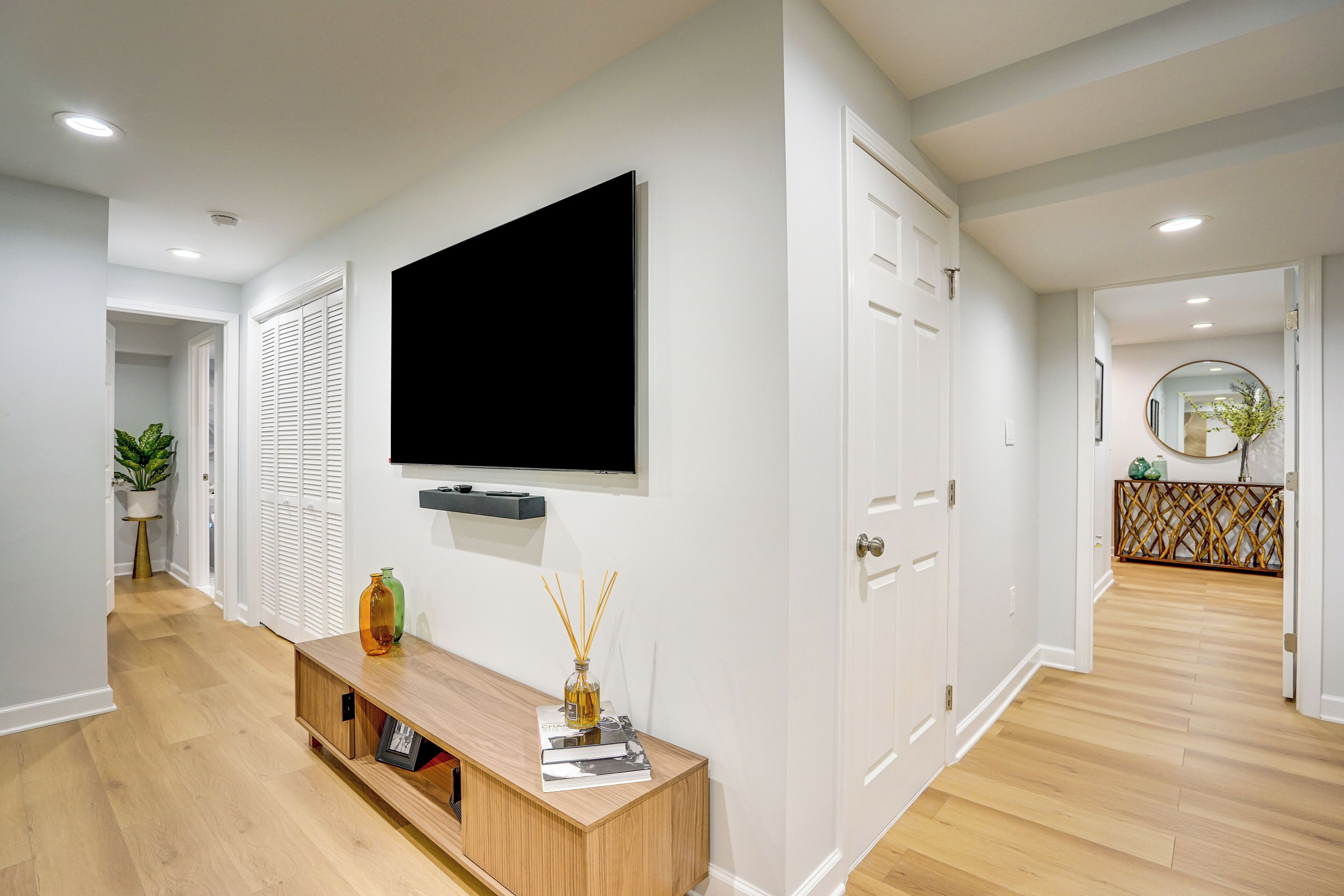 A modern hallway with a wooden floor, a mounted flat-screen TV, a console table with decorative items, and white doors.