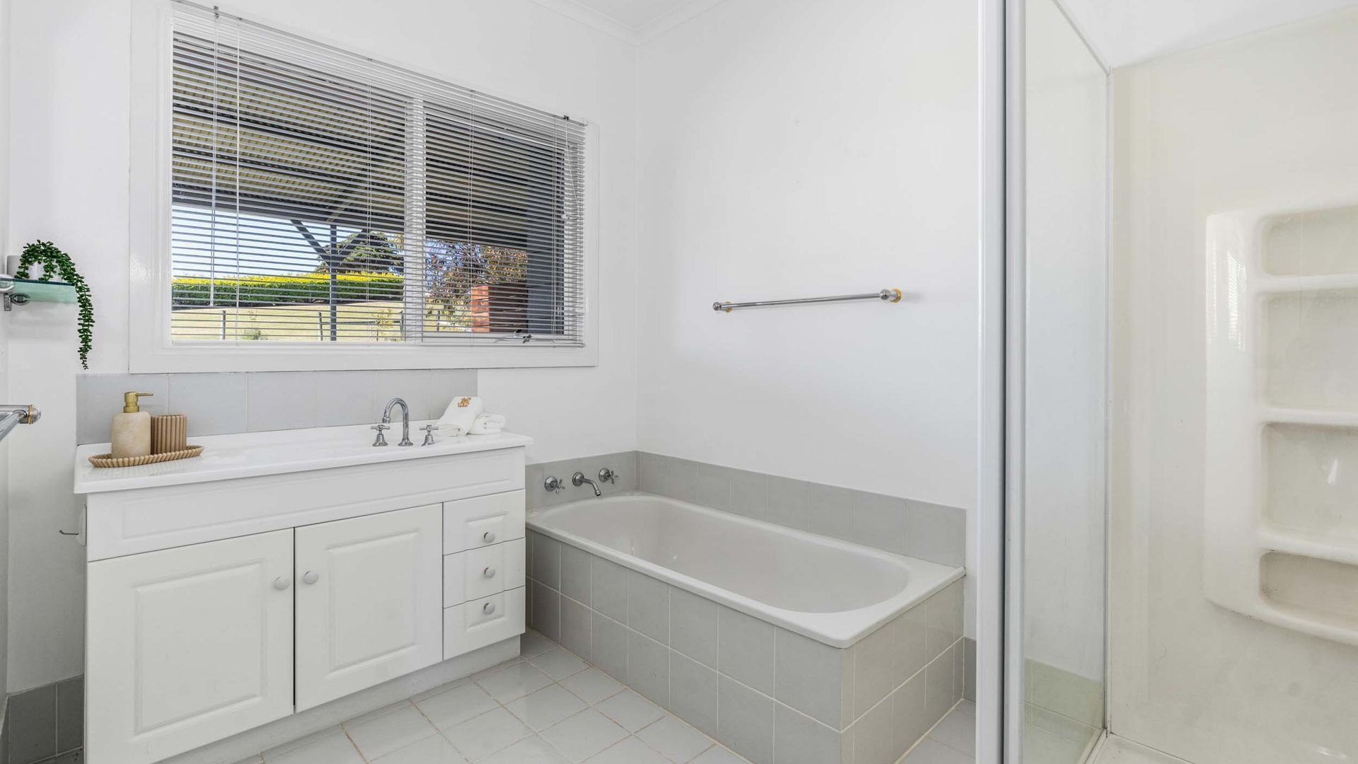A modern bathroom with a white vanity, a bathtub, and a window with blinds.