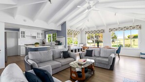 Ein großzügiges Wohnzimmer mit einer großen, grauen Eckcouch, einem Glastisch und einem Essbereich mit Holztisch und Stühlen. Im Hintergrund ist eine Küche mit weißen Schränken und einem Deckenventilator zu sehen.